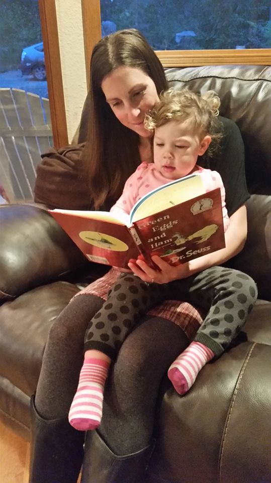 Brunette caucasian woman sitting on brown leather couch reading Green Eggs and Ham with a light brown haired caucasian toddler in lap.